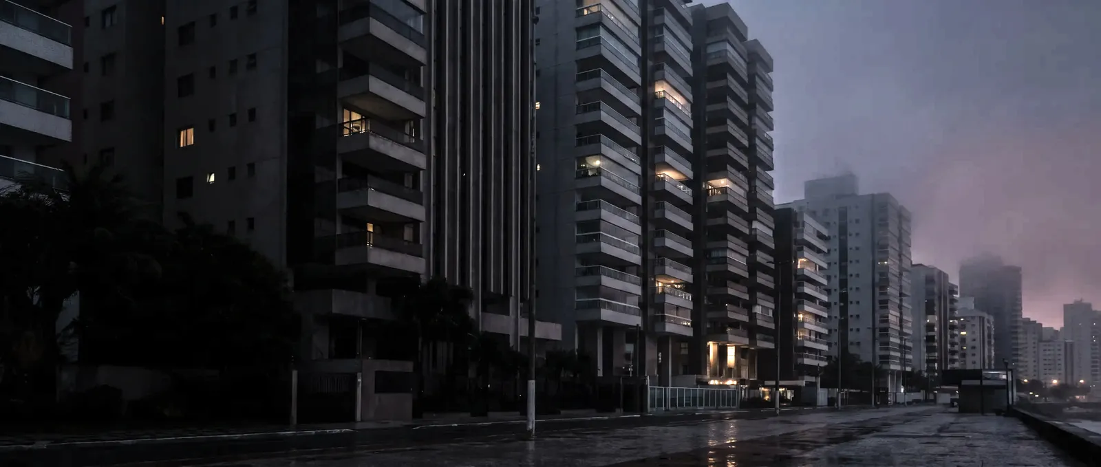Avenida de edifícios residenciais ao anoitecer, com a pista molhada refletindo as luzes